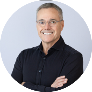 A man with short gray hair and glasses, wearing a black button-up shirt, stands smiling with his arms crossed in front of a light background.