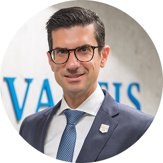 Man in a suit and tie with glasses, standing indoors in front of a blurred sign with blue letters.