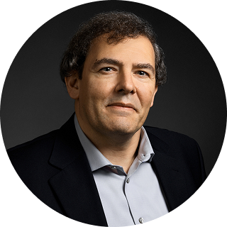 A middle-aged man with curly dark hair wearing a black blazer and light gray shirt poses against a dark background.