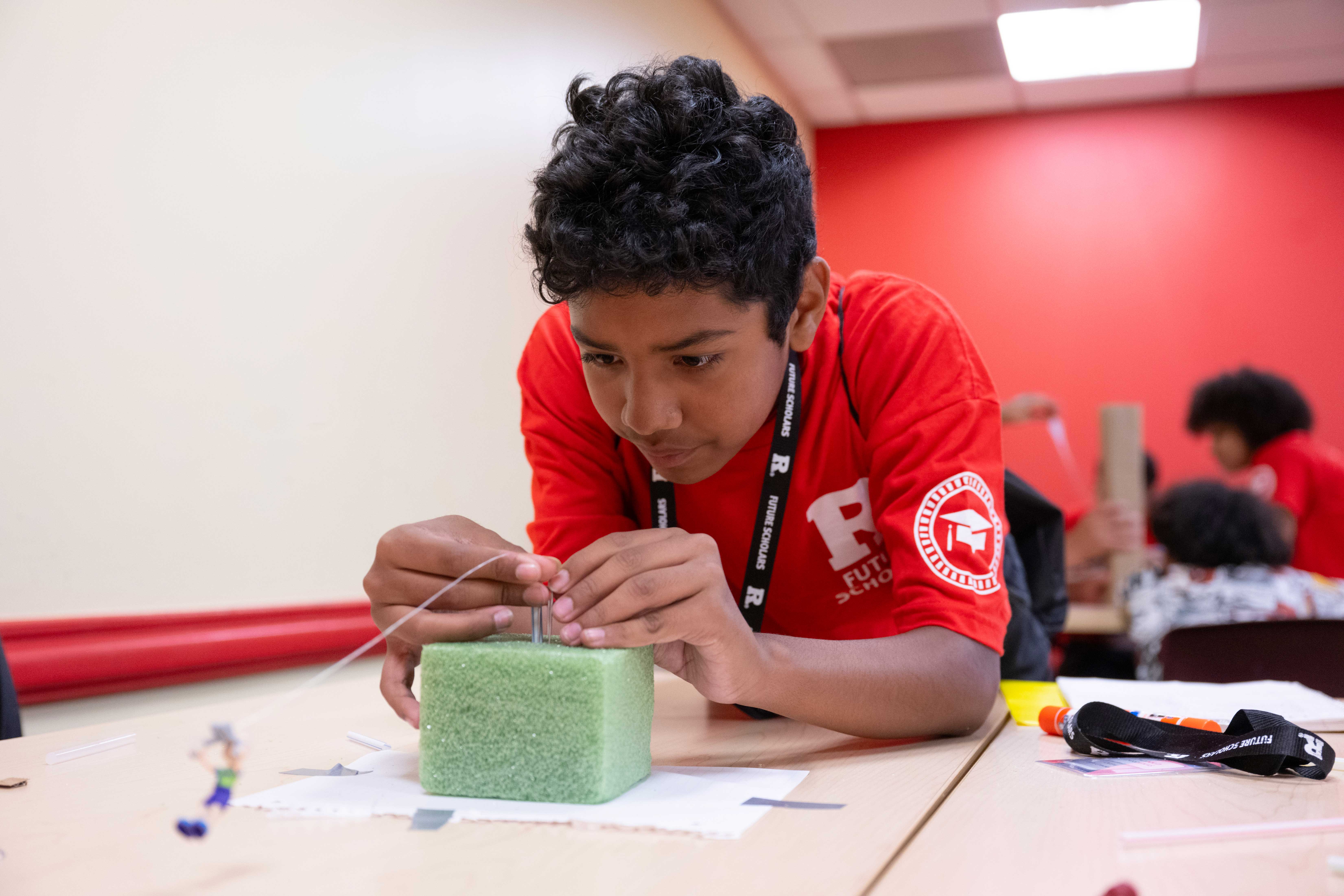 A boy in a red shirt concentrates while building a small project using string and a green foam block at a classroom table.