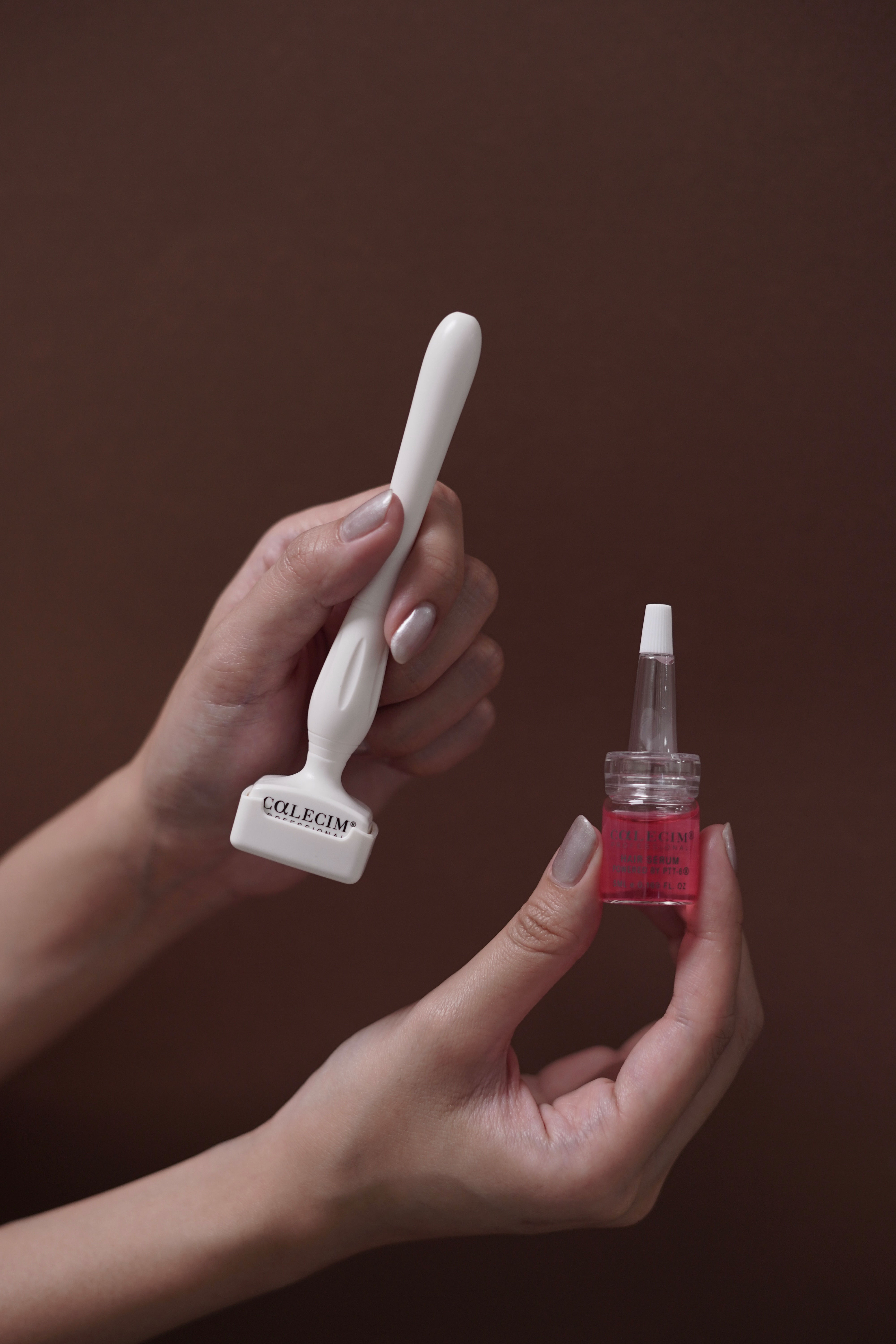 A person holds a white derma roller in one hand and a small bottle of pink serum with a dropper in the other, against a brown background.