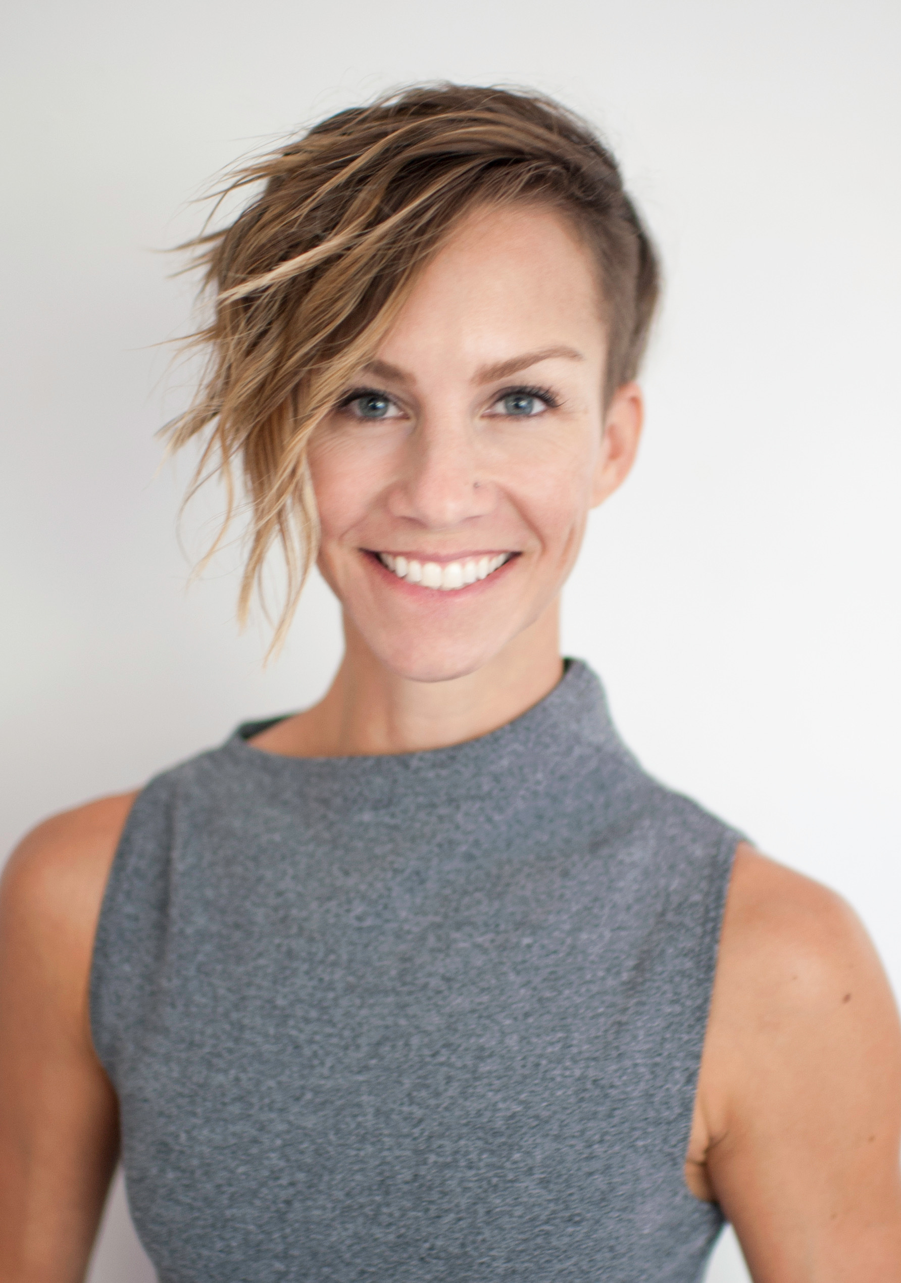 A woman with short, asymmetrical blonde hair and a sleeveless gray top smiles at the camera against a plain white background.