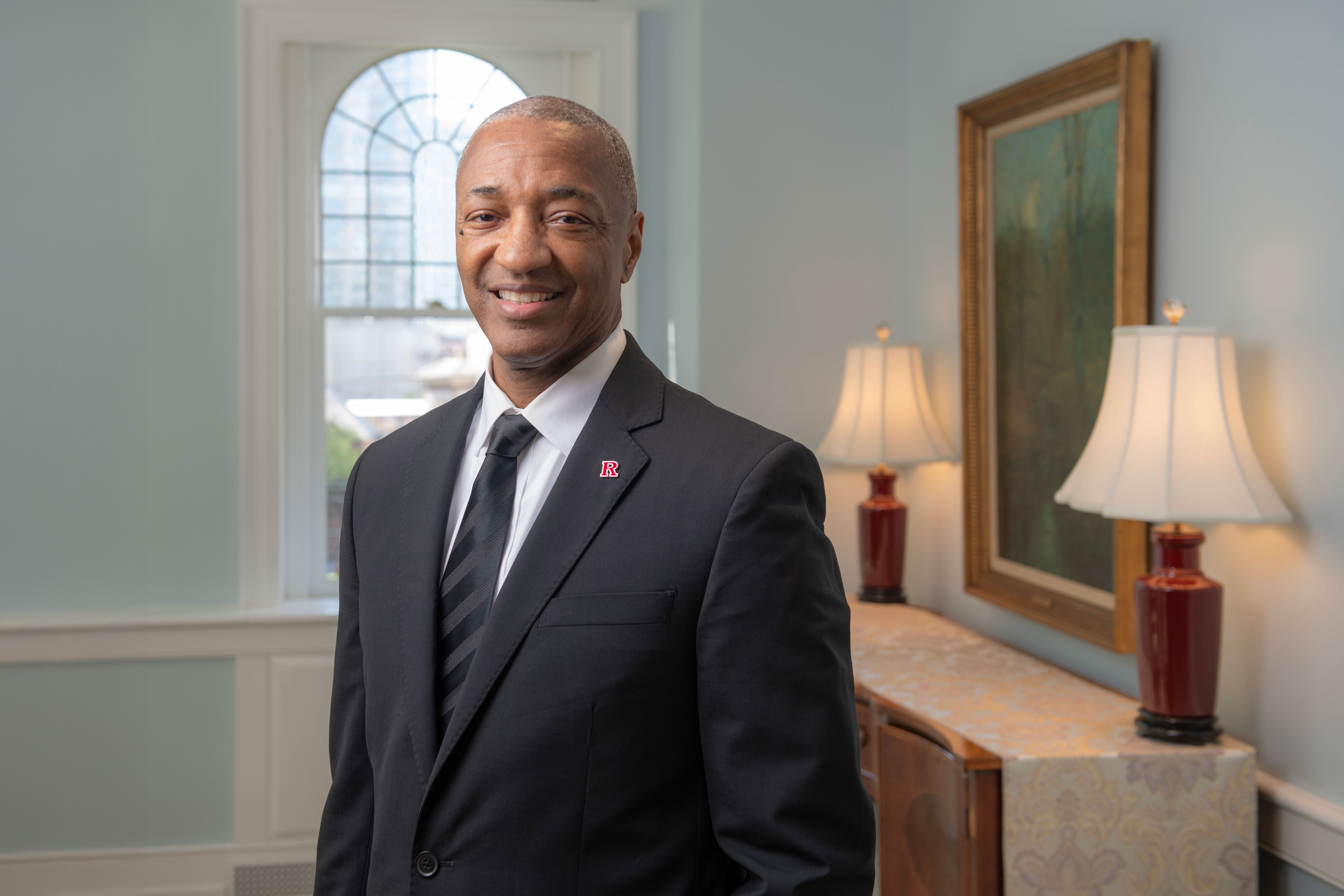 A man in a suit and tie stands in a well-lit room with two lamps, a framed painting, and a window in the background.
