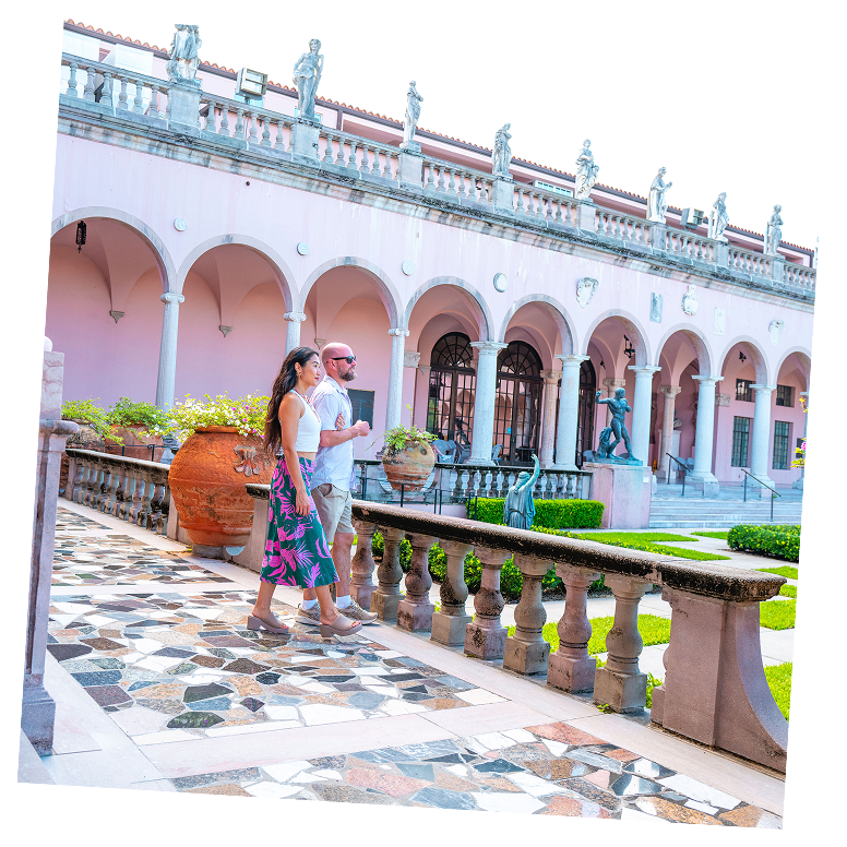 A man and woman walk along a tiled terrace in front of a pink building with arches and statues, surrounded by a manicured garden.