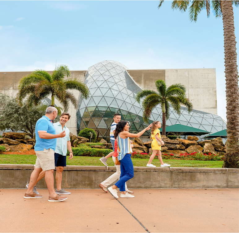 A group of six people walk outside a modern glass and concrete building surrounded by palm trees and landscaping on a sunny day.