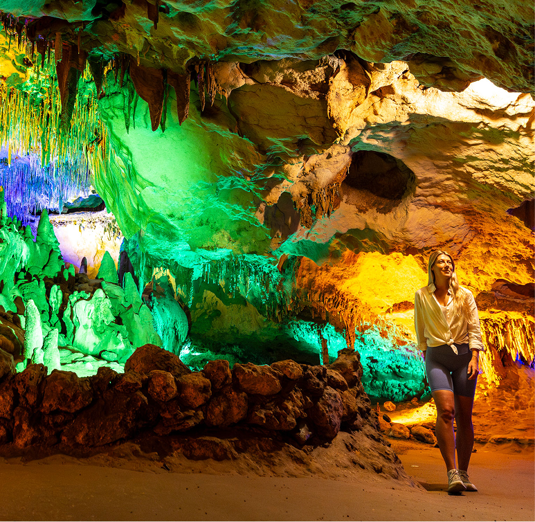 A person walks inside a cave illuminated by colorful lights, highlighting rock formations and stalactites on the ceiling.