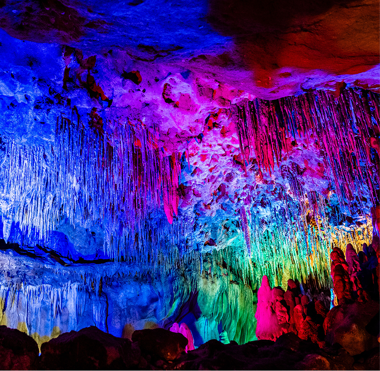 Colorful artificial lights illuminate stalactites and stalagmites inside a cave, highlighting the natural rock formations in vibrant shades of blue, pink, red, and green.