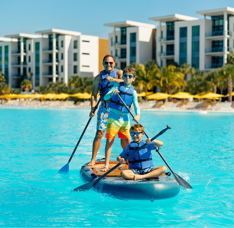 Three people in life jackets paddle boarding on a bright blue pool, with modern buildings and yellow umbrellas in the background.