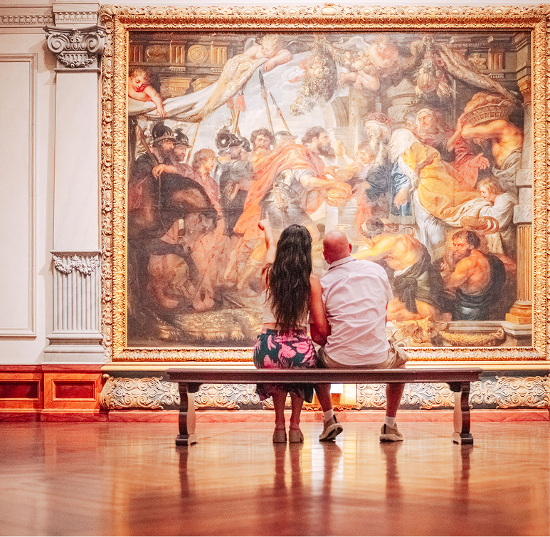 A man and woman sit on a bench in an art gallery, viewing a large, detailed classical painting in an ornate gold frame.