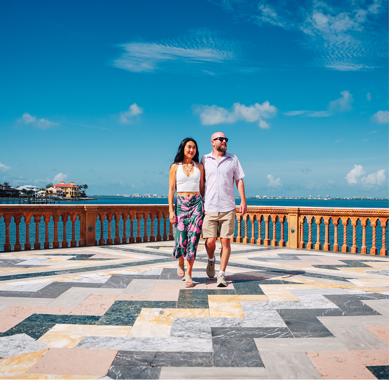A woman and a man walk side by side on a patterned terrace with a waterfront and blue sky in the background.