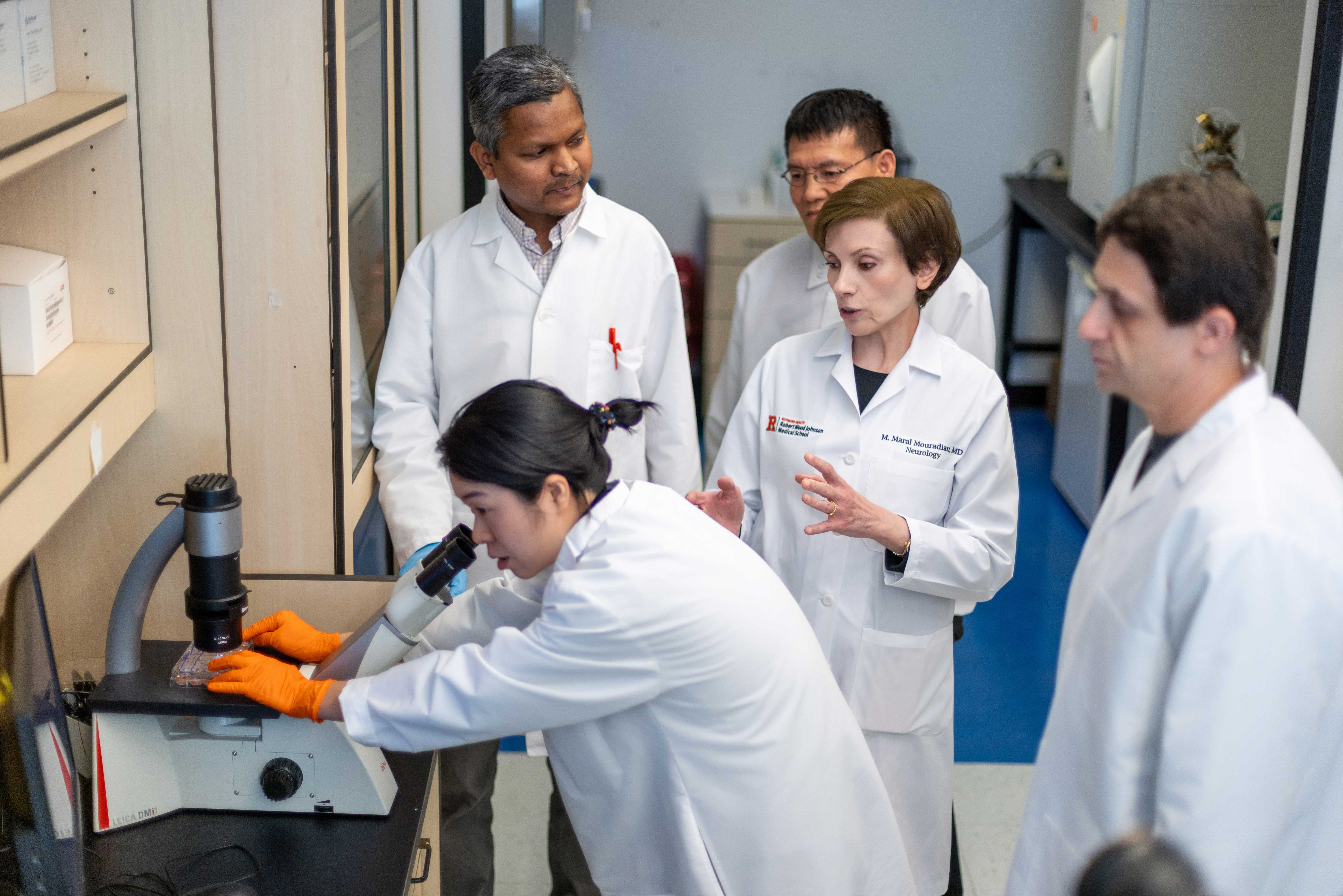 Five scientists in lab coats are gathered in a laboratory; one is looking through a microscope while the others observe and discuss.