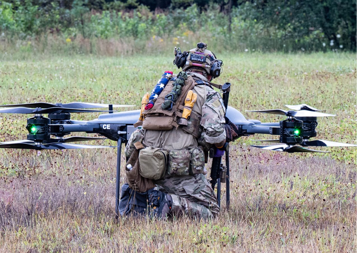 A soldier in camouflage gear kneels beside a large drone in a grassy field, holding a rifle and wearing a helmet with tactical equipment.
