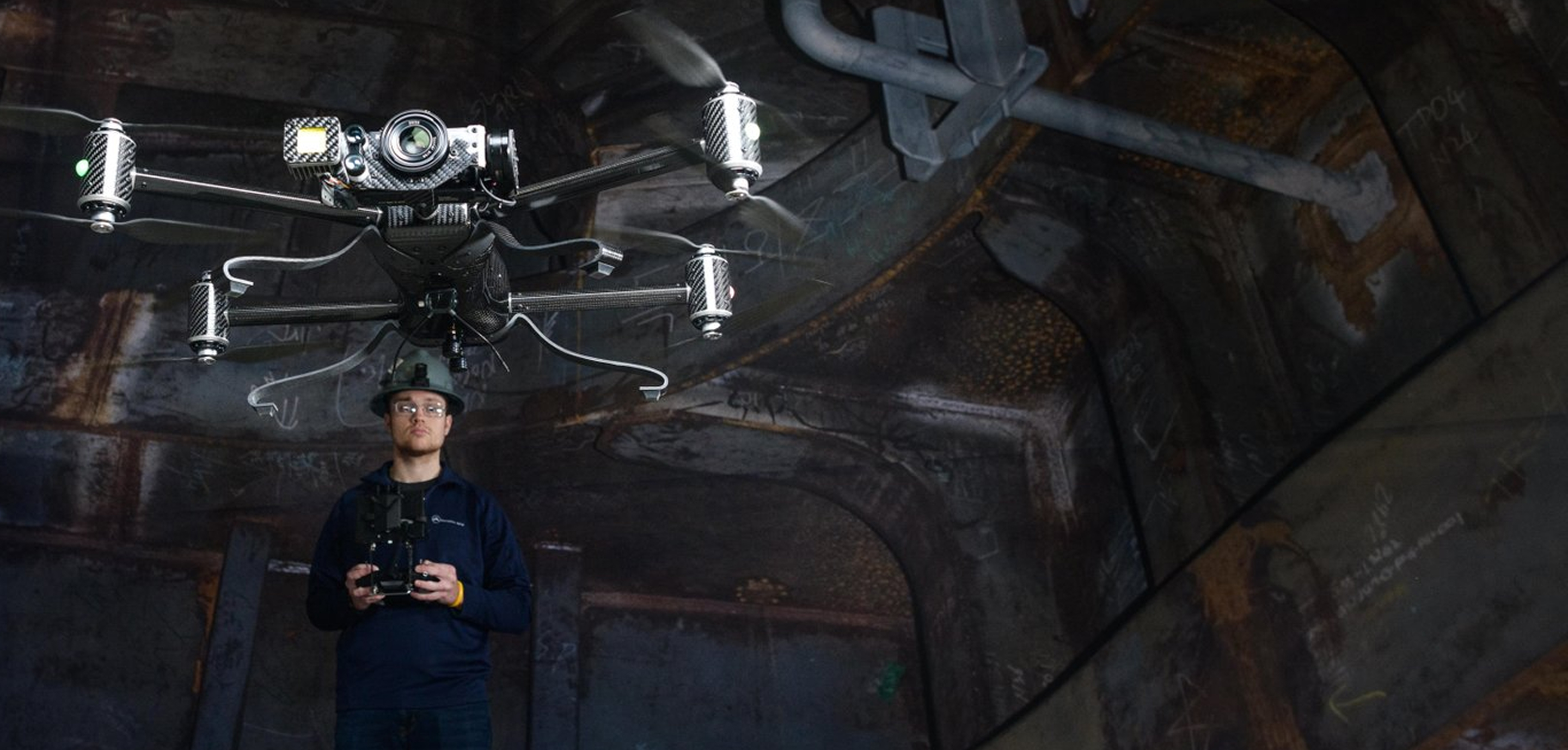 A person wearing safety glasses operates a large drone inside a dimly lit industrial facility with metal walls and pipes.
