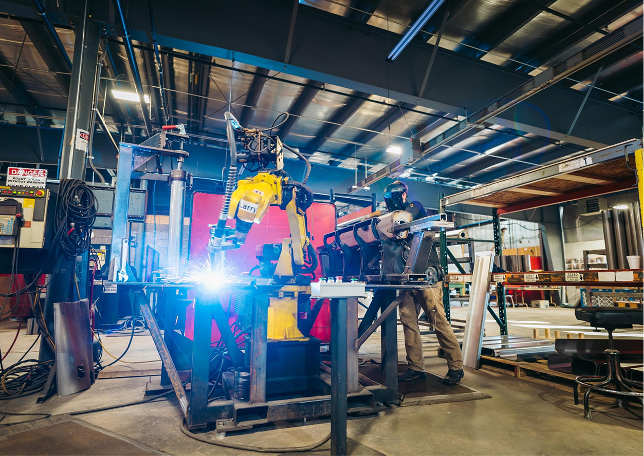 A robotic arm performs automated welding on metal parts in an industrial factory, with a worker monitoring the process nearby.