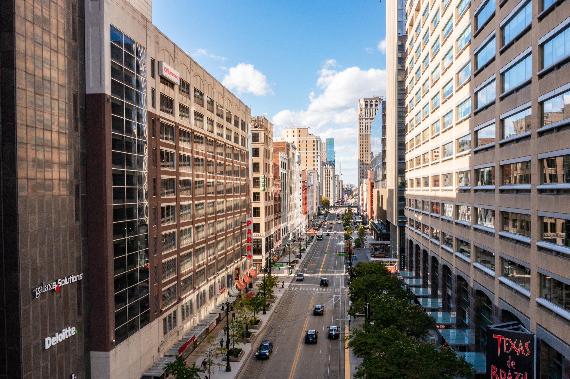A wide city street runs between tall office buildings under a partly cloudy sky, with cars and pedestrians visible on the road and sidewalks.
