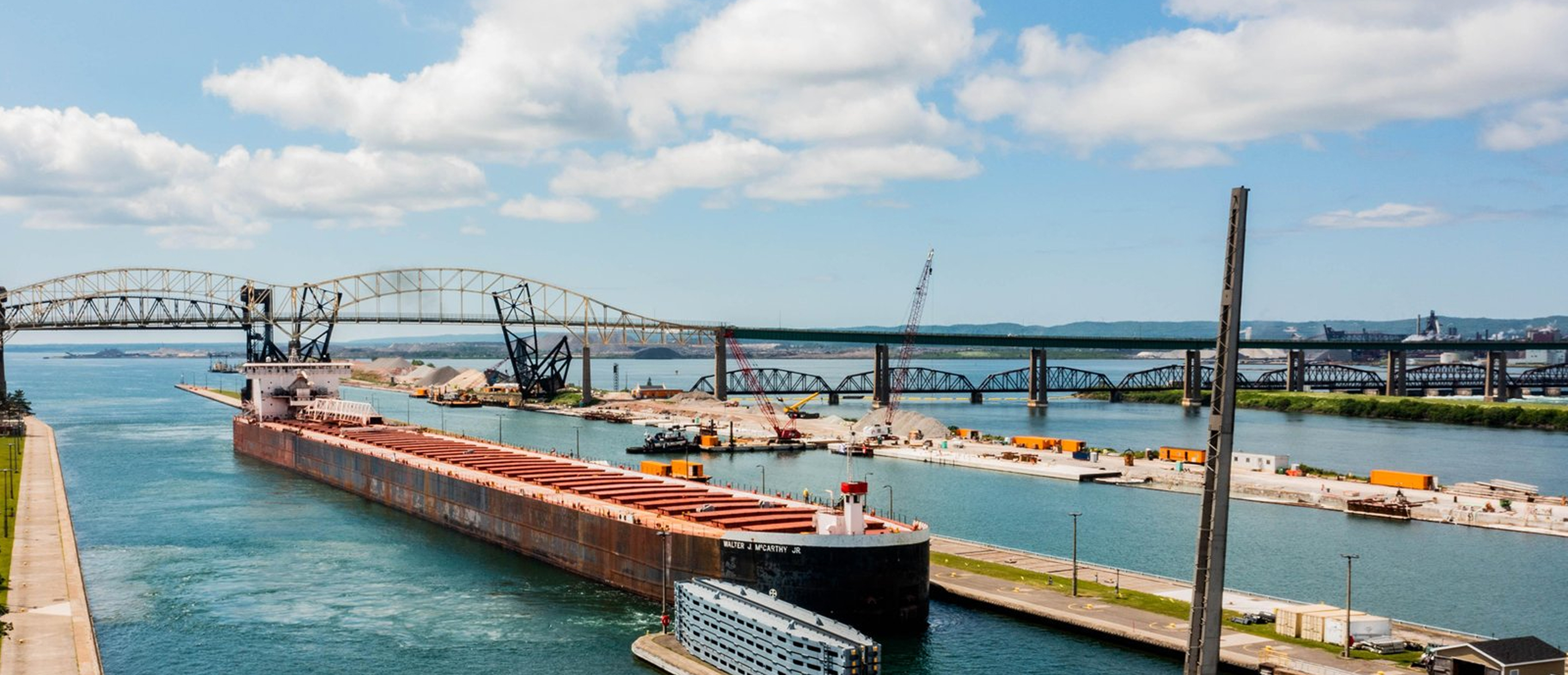 A large cargo ship passes through a canal with bridges in the background, construction equipment, and blue sky with clouds overhead.