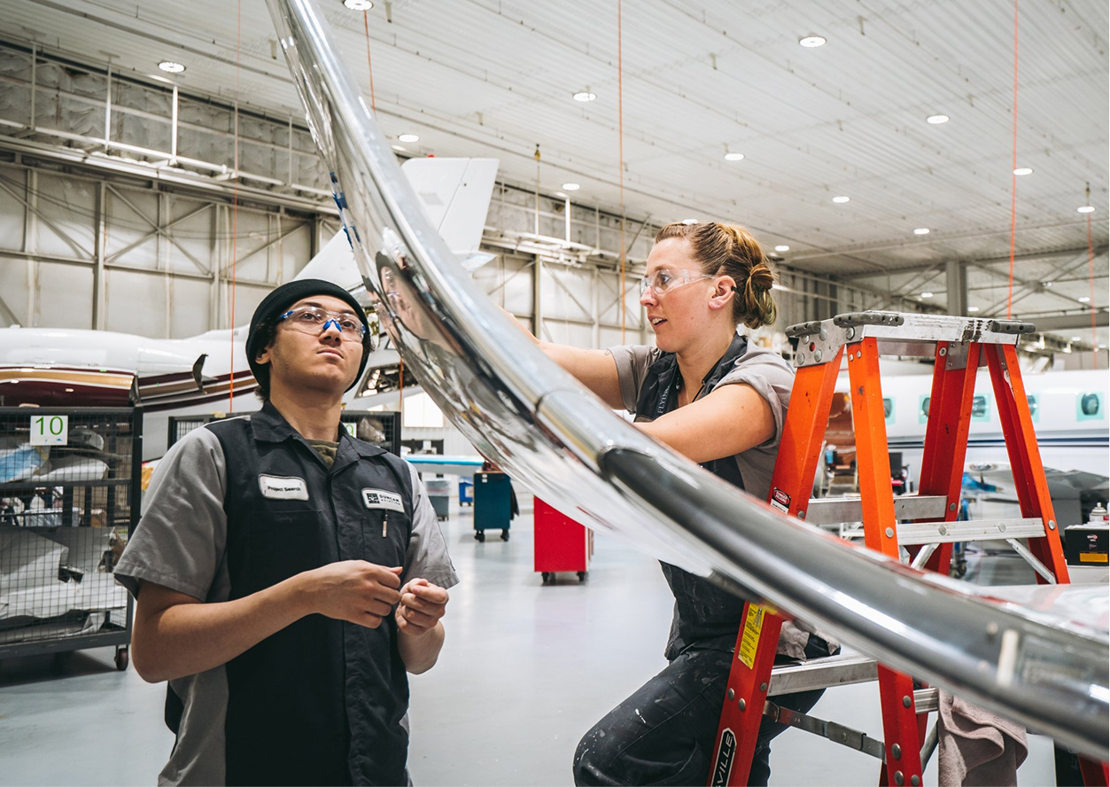 Two aviation technicians work on the wing of an aircraft inside a hangar; one stands on a ladder while the other observes from the ground.