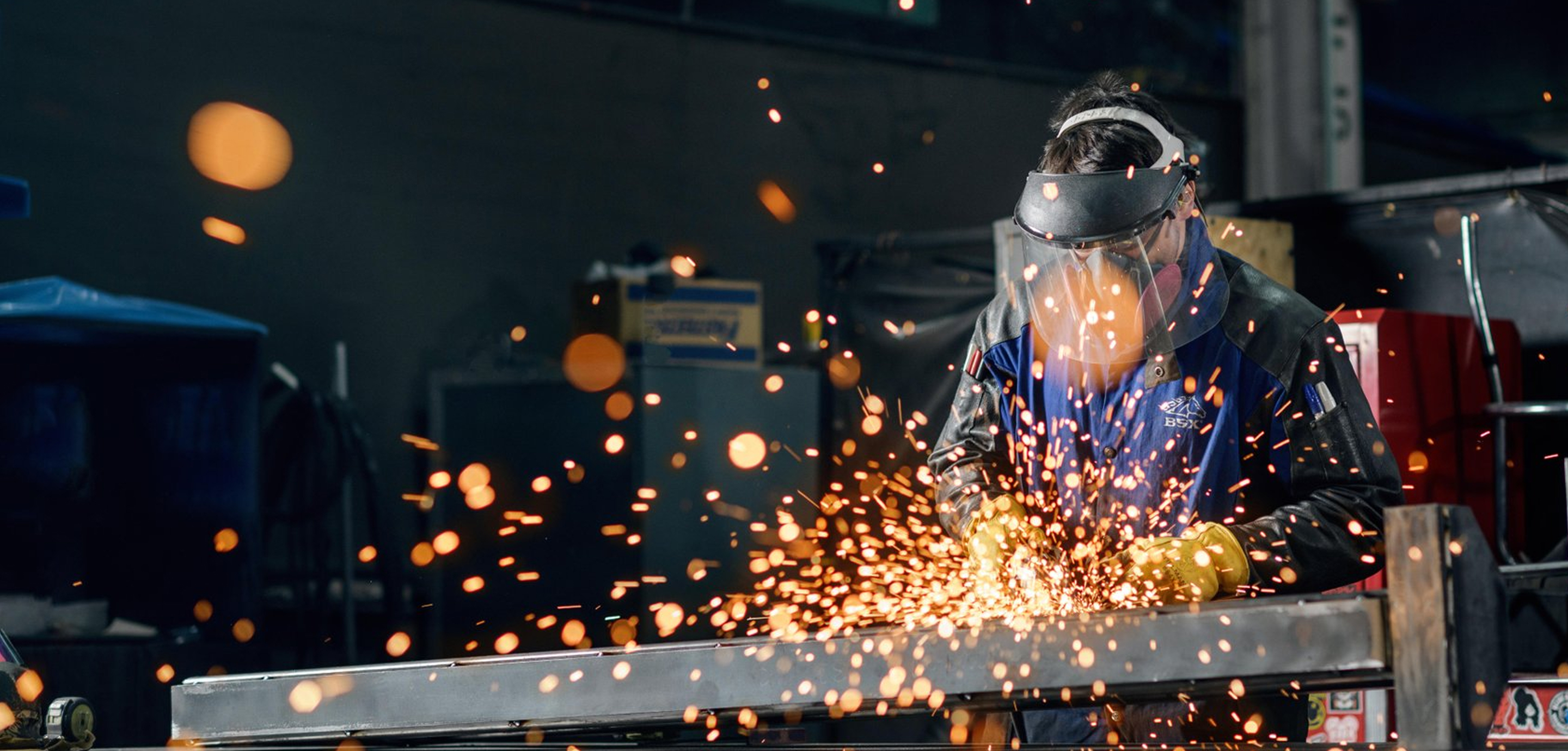 A person wearing safety gear uses a power tool to grind metal, producing sparks in an industrial workshop setting.