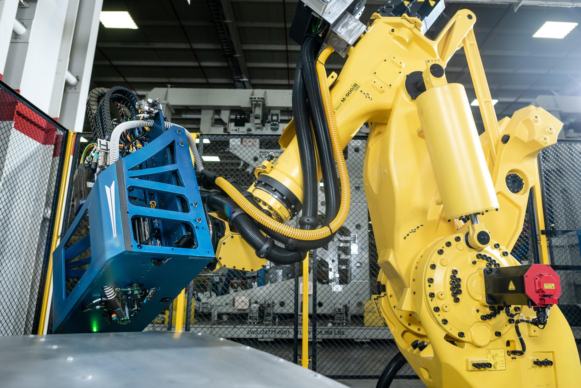 A yellow industrial robotic arm operates a blue mechanical component in a factory setting, with safety fencing and machinery visible in the background.