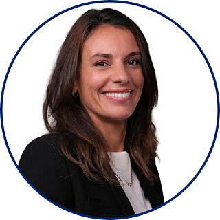 Woman with long brown hair wearing a black blazer and white top, smiling at the camera against a plain white background with a blue circular border.