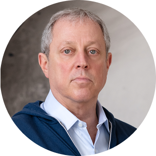 A middle-aged man with short gray hair wearing a light blue shirt and a dark jacket, looking directly at the camera against a neutral background.