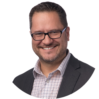 Man with short brown hair, glasses, and beard, wearing a dark blazer over a checked shirt, smiling at the camera against a plain white background.