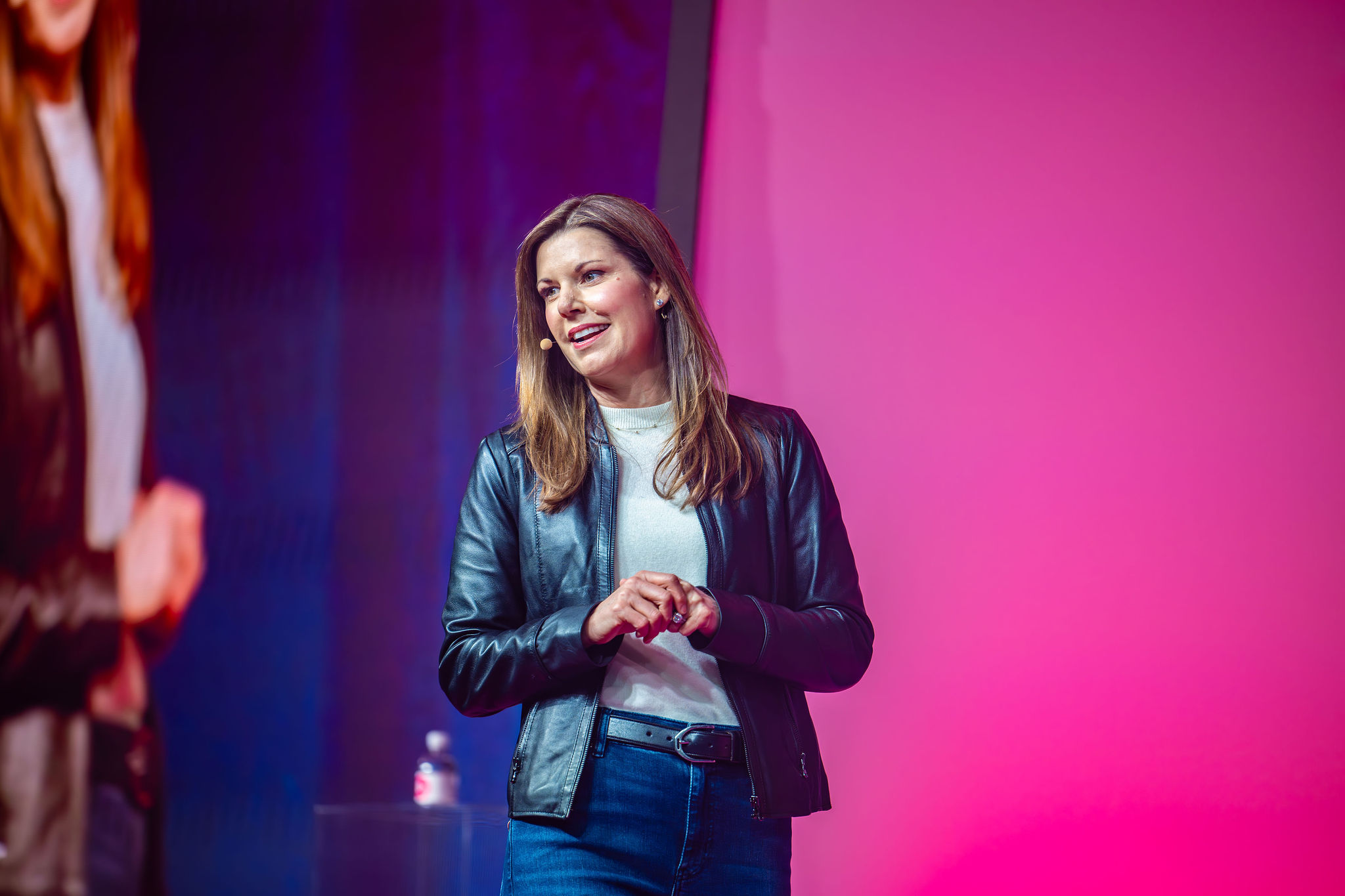 A woman wearing a black leather jacket and jeans stands on stage, speaking, with a pink and blue background behind her.