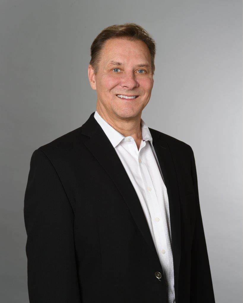 A man in a black suit jacket and white dress shirt stands in front of a plain gray background, smiling at the camera.