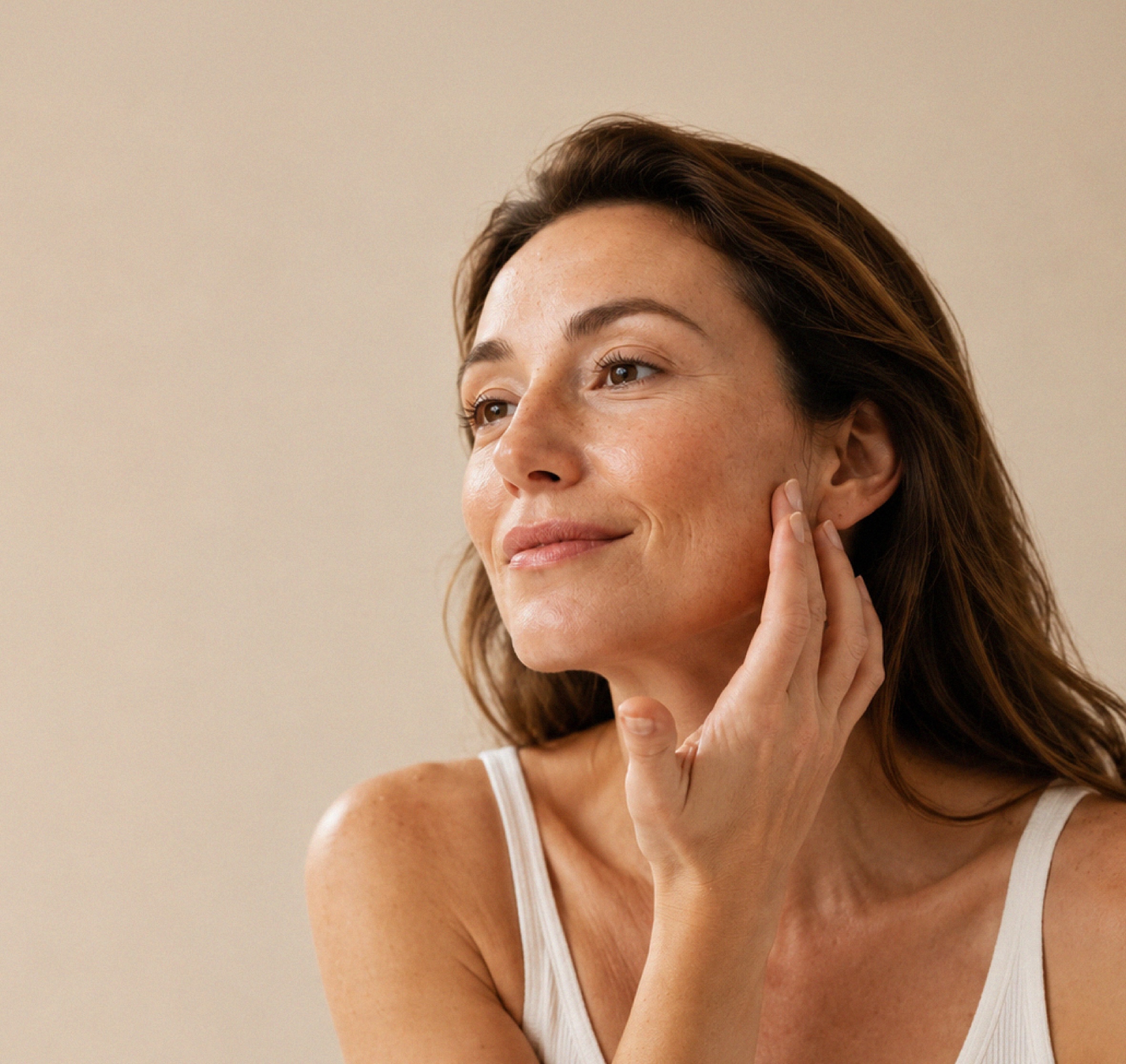 Woman with long brown hair in a white tank top touching her face gently and looking slightly to the side, against a plain beige background.