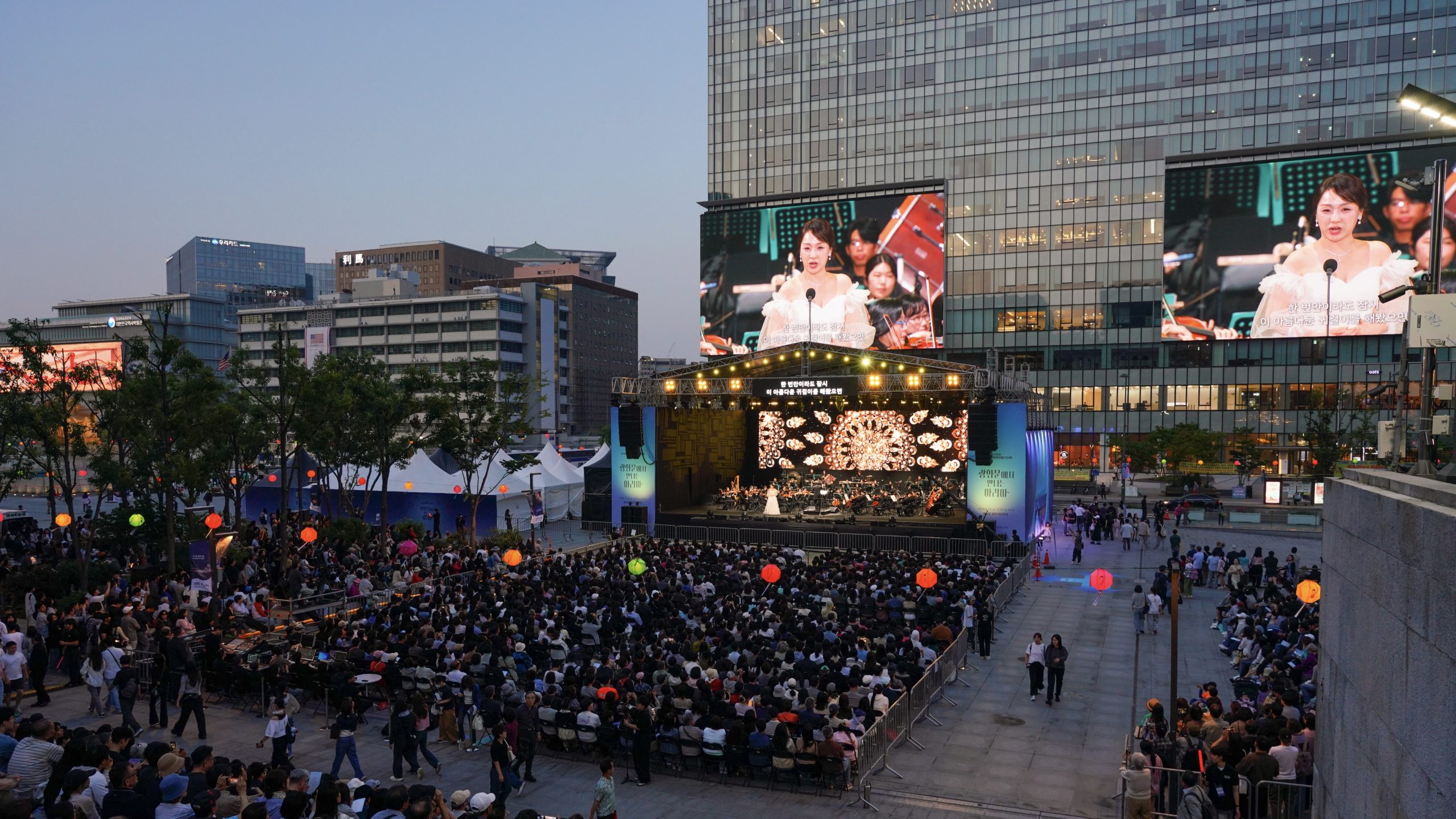A large audience watches an outdoor concert on a stage with screens showing a woman speaking, set in an urban plaza with tall buildings in the background.