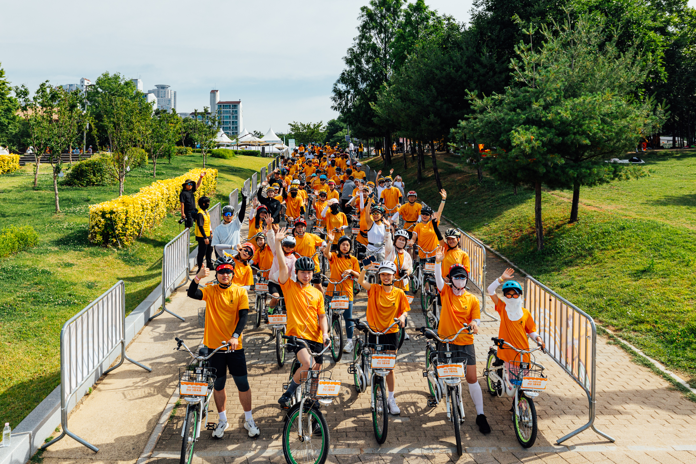 A large group of people in orange shirts ride bicycles together along a paved path, surrounded by greenery and metal barriers, on a sunny day.