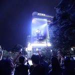 People take photos with phones of a large digital outdoor screen displaying a musician at night in an urban setting with trees and a tall building in the background.