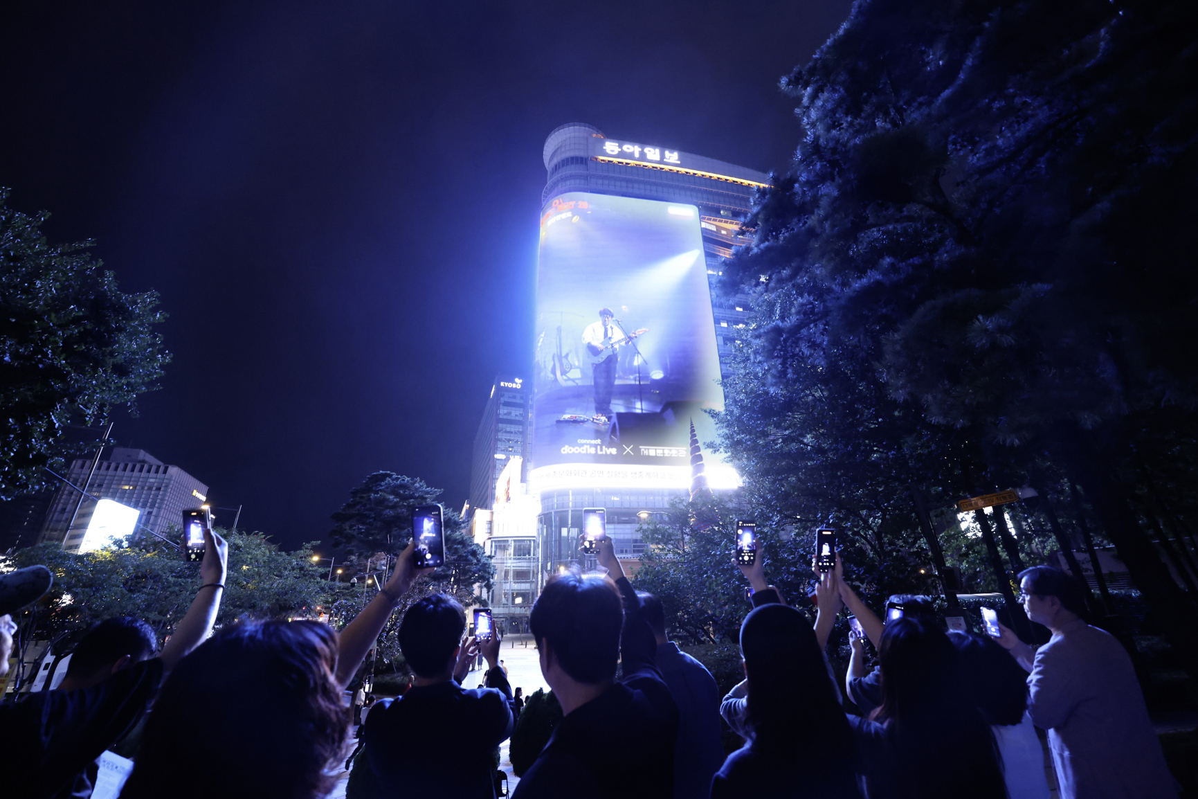 People take photos with phones of a large digital outdoor screen displaying a musician at night in an urban setting with trees and a tall building in the background.